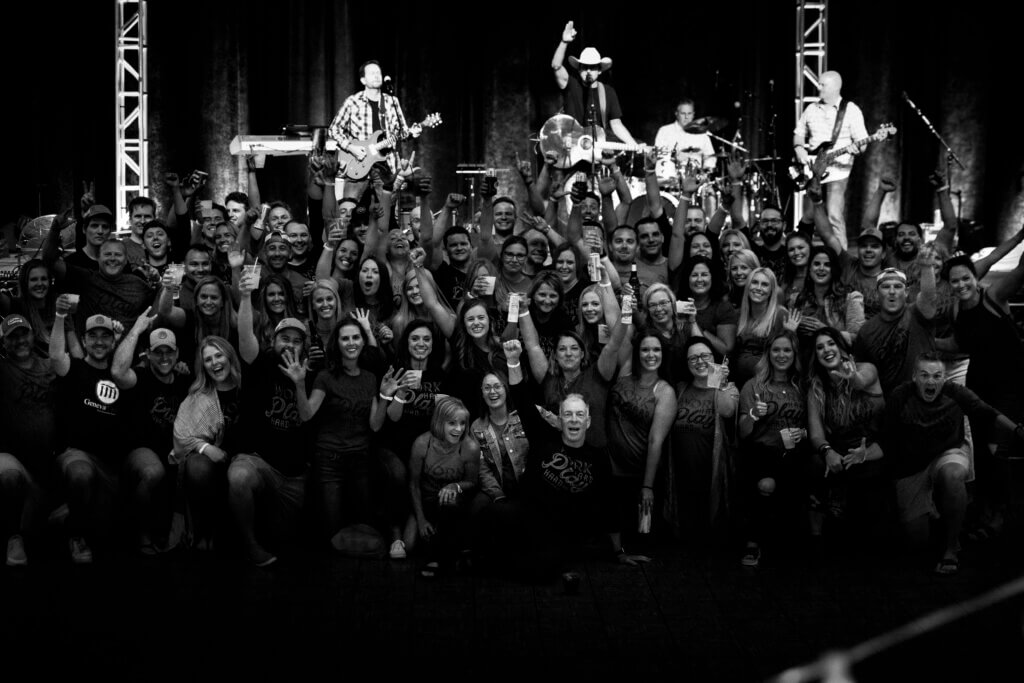 Large group of team members posing together at concert venue with band performing on stage behind them.