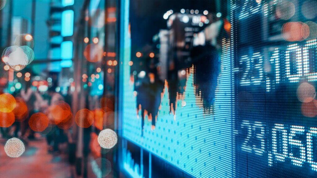 Blue LED financial display showing stock charts and economic data with blurred city lights in background.