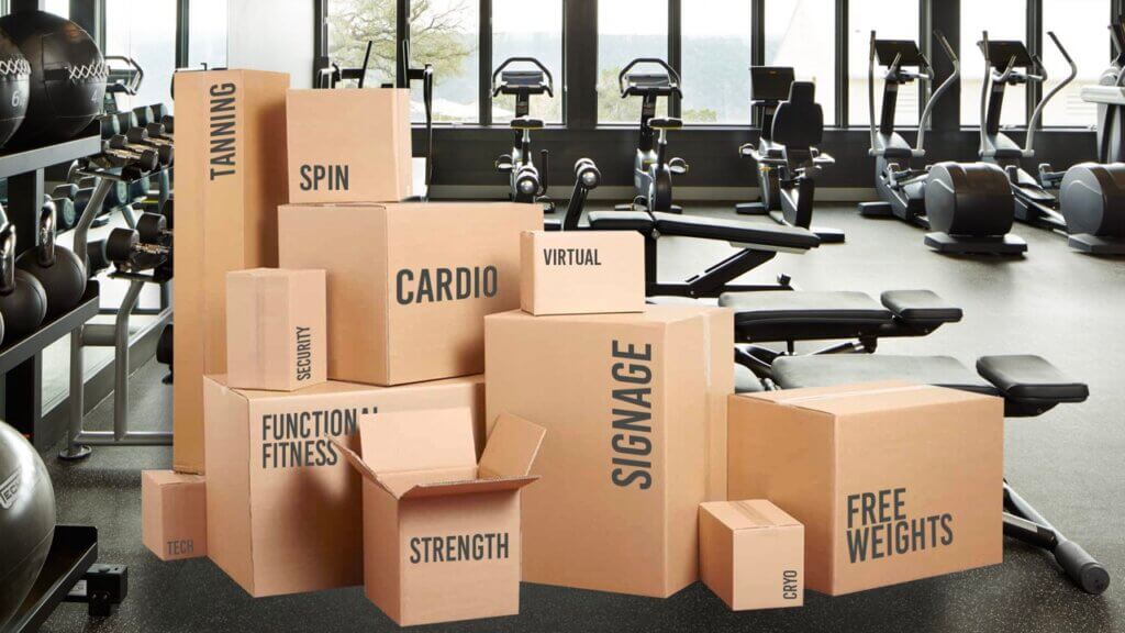 Cardboard boxes with fitness labels like "CARDIO" and "STRENGTH" stacked in gym with exercise equipment.