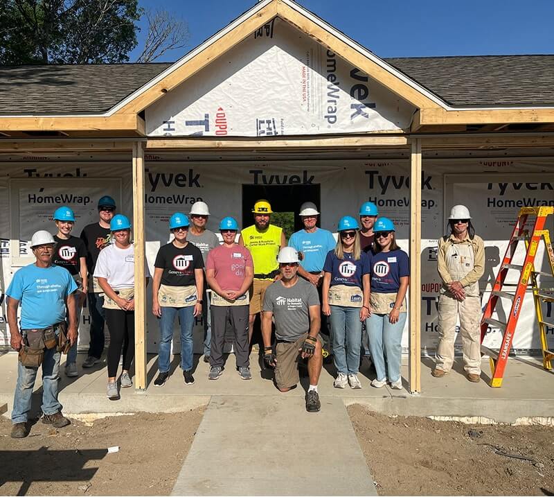 Fourteen volunteers in hard hats and work clothing standing together at residential construction site with house frame behind them.