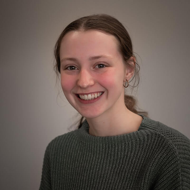 Young woman with brown hair pulled back smiling in olive green knit sweater against gray background