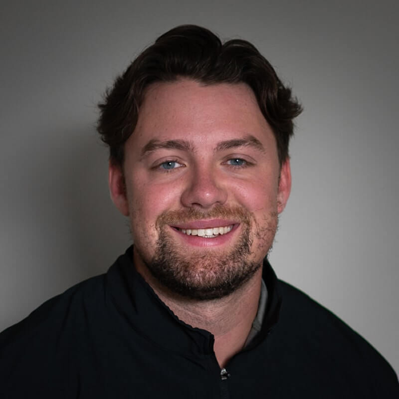 Man with dark wavy hair and beard smiling in black polo shirt against gray background