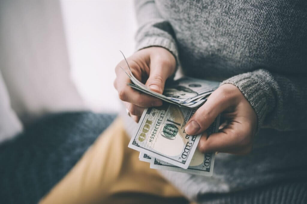 Hands holding stack of one hundred dollar bills being counted with person wearing gray sweater.
