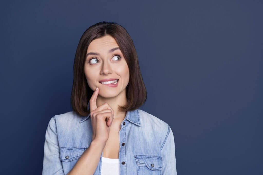 Woman with brown bob haircut wearing denim shirt looking up thoughtfully with finger to chin against blue background.