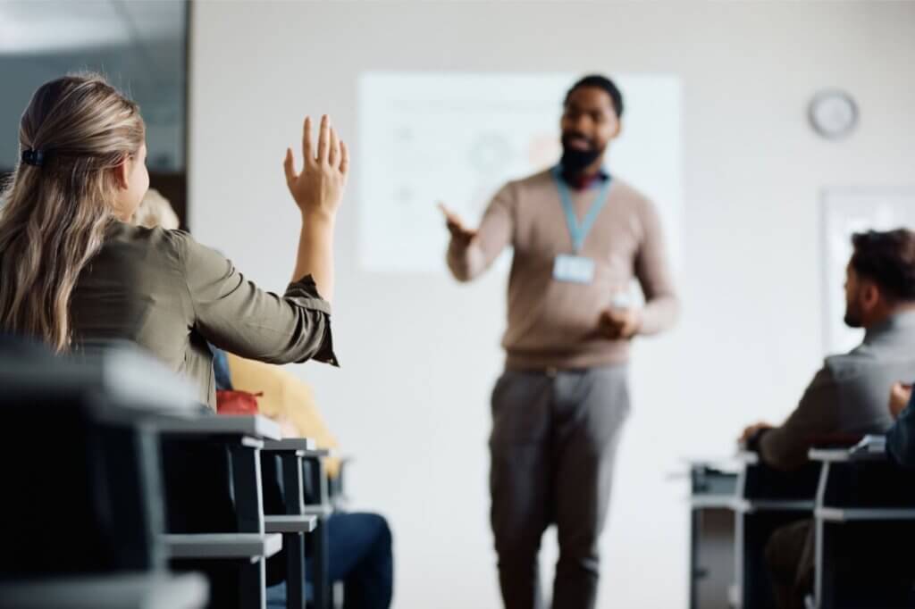 Woman in business attire raising hand during conference presentation with presenter visible in background at whiteboard.