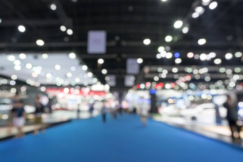 Blurred image of convention center exhibition floor with blue carpet, booth displays, and overhead lighting creating bokeh effect.