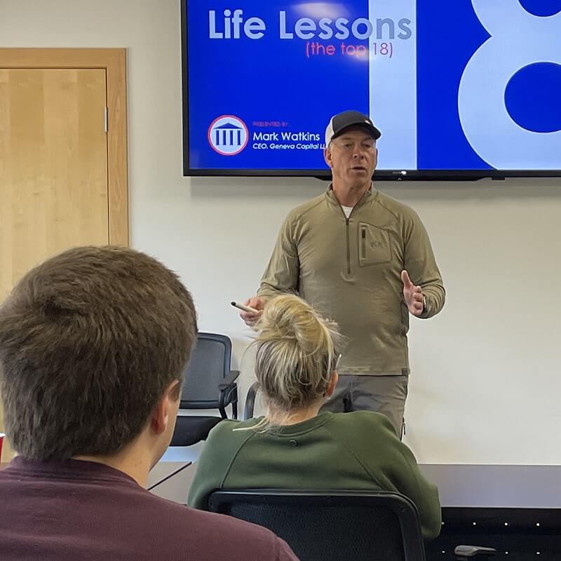 Man in casual attire presenting to seated audience with Life Lessons presentation visible on blue screen behind him.