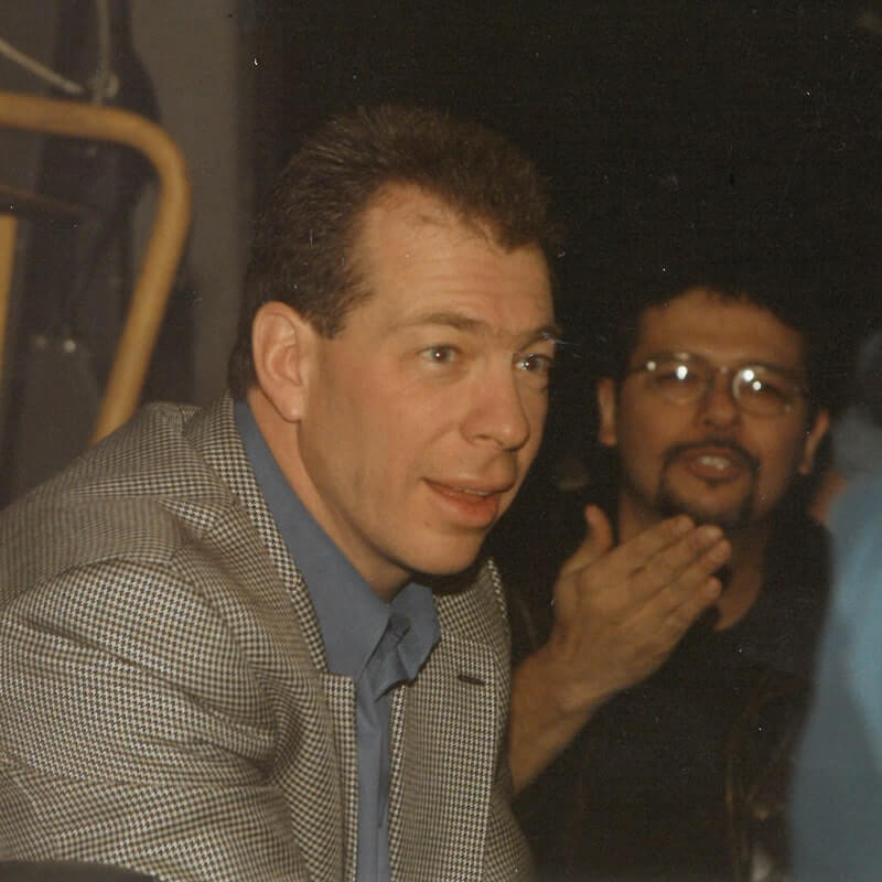Vintage photo of man in checkered suit jacket with another person visible in background wearing glasses.