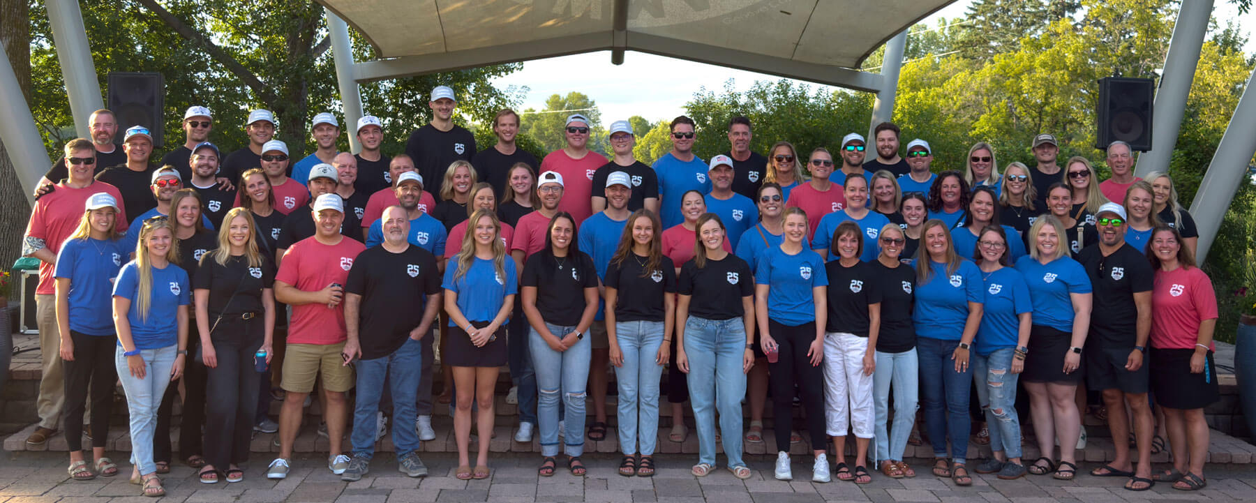 Large group of company employees in branded shirts standing together for team photo under outdoor pavilion with trees visible in background.