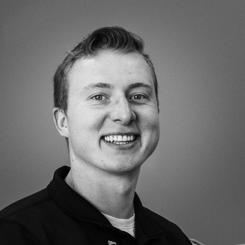 Black and white photo of a young, brown-haired man with a black polo shirt on and smiling.