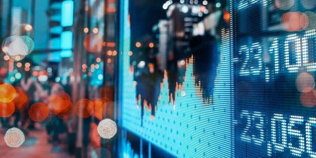 Blue LED financial display showing stock charts and economic data with blurred city lights in background.