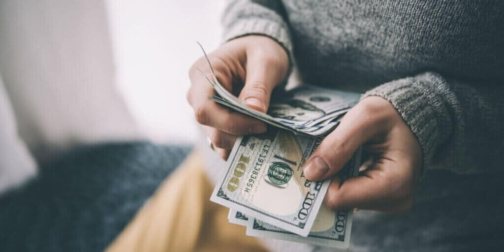 Hands holding stack of one hundred dollar bills being counted with person wearing gray sweater.