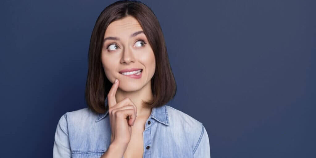 Woman with brown bob haircut wearing denim shirt looking up thoughtfully with finger to chin against blue background.