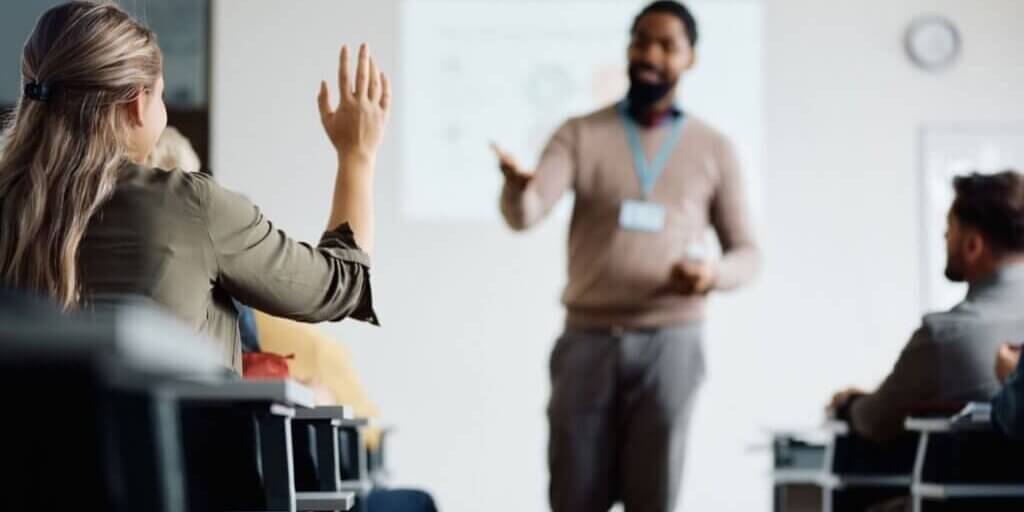 Woman in business attire raising hand during conference presentation with presenter visible in background at whiteboard.