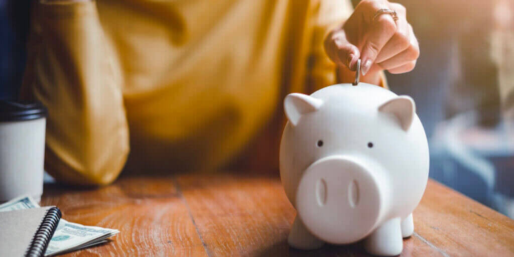 Hand inserting coin into white piggy bank sitting on wood surface next to dollar bills and spiral notebook.