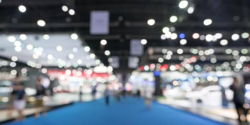 Blurred image of convention center exhibition floor with blue carpet, booth displays, and overhead lighting creating bokeh effect.