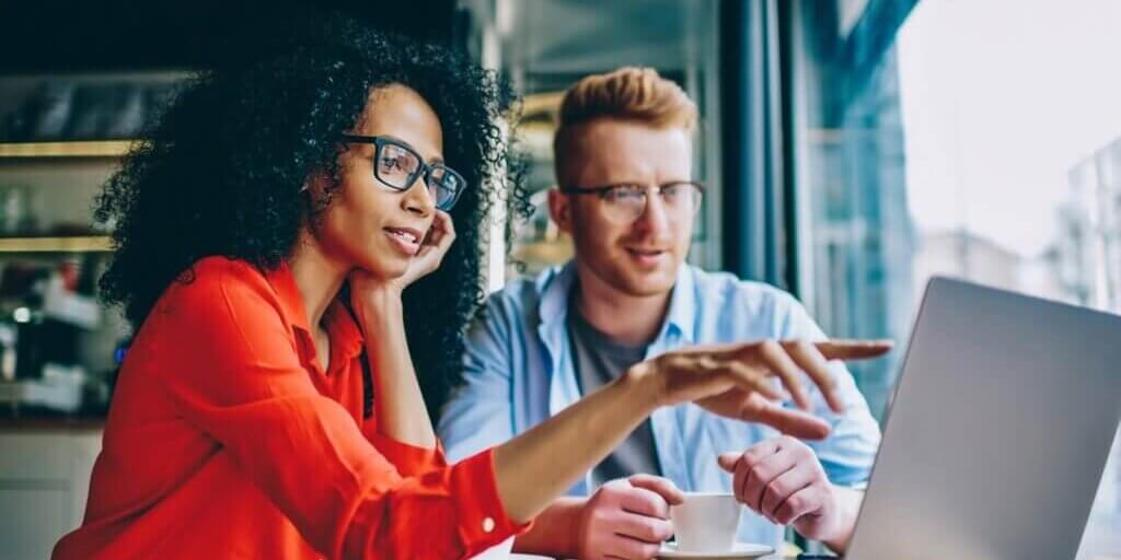 Woman with curly hair wearing red shirt and glasses pointing at laptop screen while collaborating with male colleague in blue shirt.