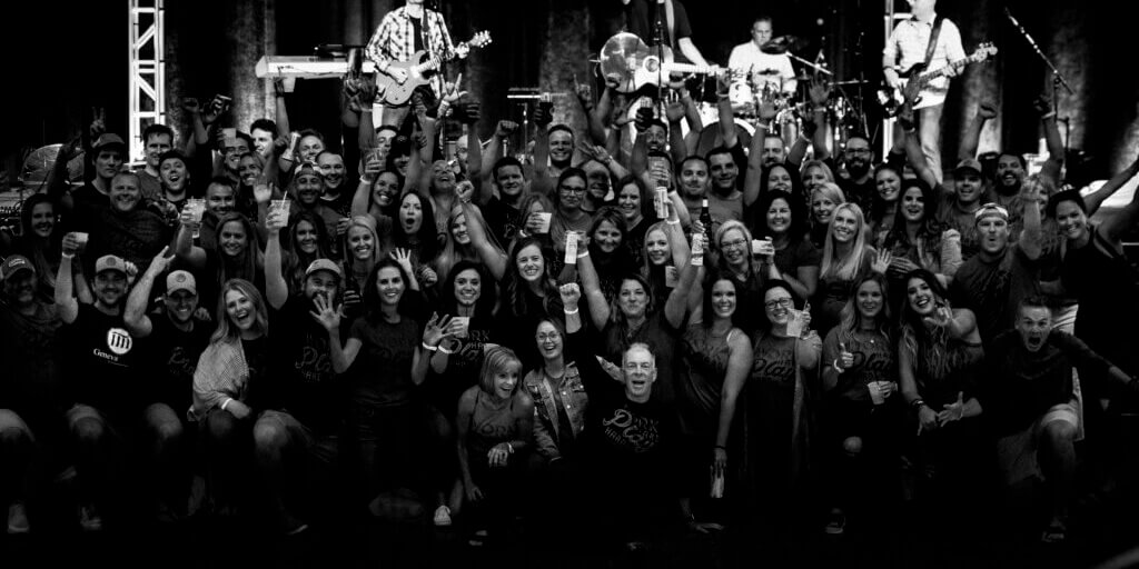 Large group of team members posing together at concert venue with band performing on stage behind them.