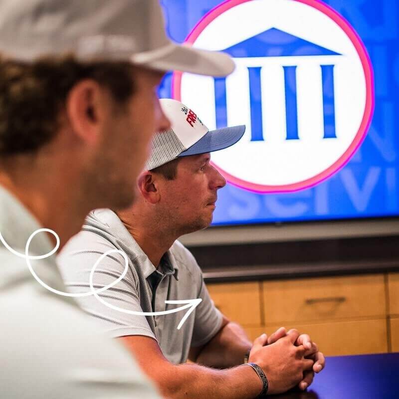 Two men in polo shirts and baseball caps sitting at meeting table with large screen showing circular logo with classical building columns in background.