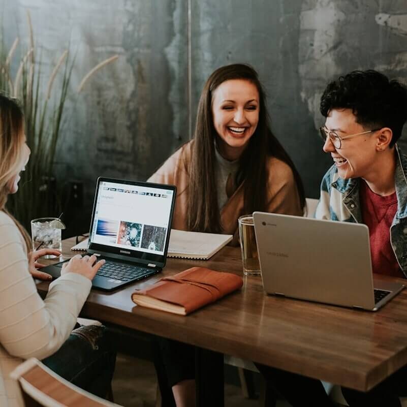 Three people smiling and working on laptops at wooden table in modern office with exposed brick wall.