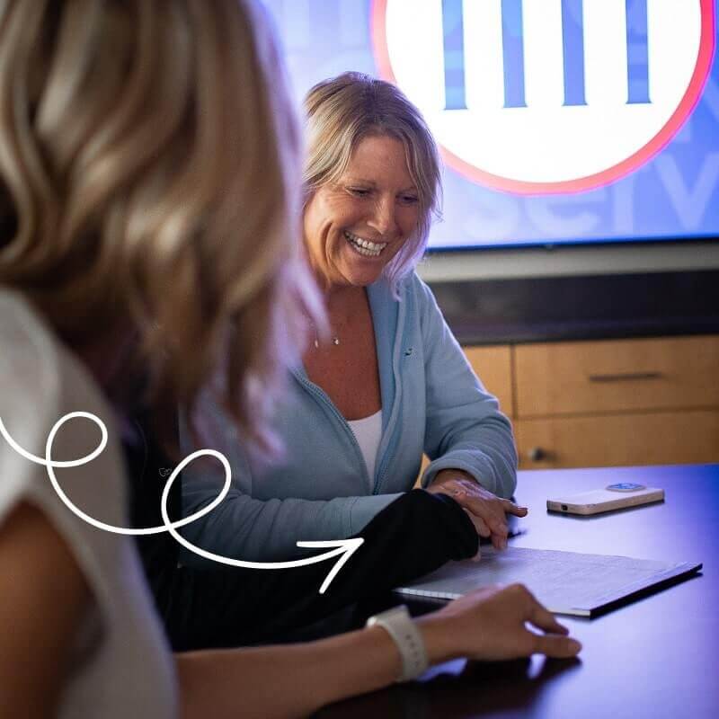Woman with blonde hair in blue blazer smiling during business meeting at conference table with institutional logo visible on screen behind her.