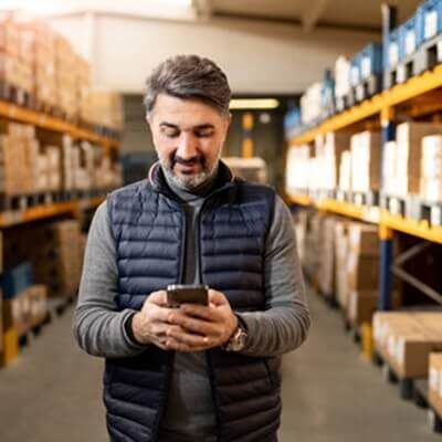 Smiling man in dark vest using phone in warehouse with tall storage shelves and boxes in background.