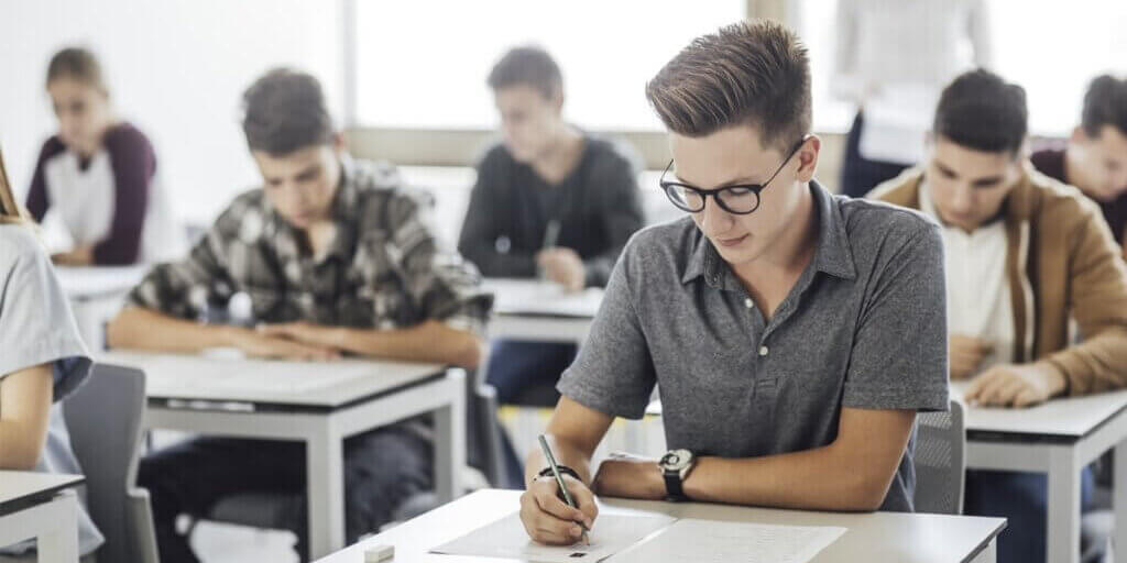 Student in glasses writing exam with pen while classmates work at desks in bright classroom.