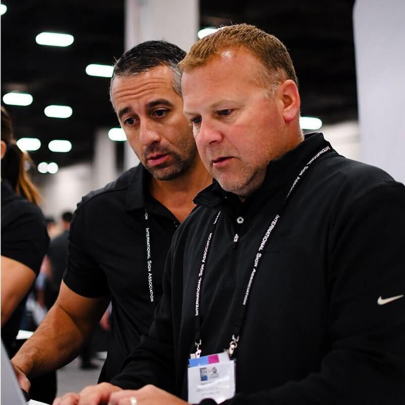 Two men in black shirts with conference badges looking at laptop screen together in convention center with overhead lighting visible.