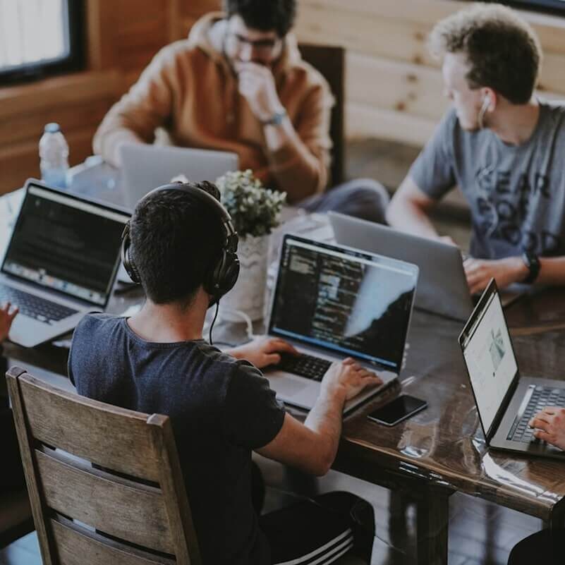 Three people working on laptops, one person wearing headphones, in collaborative workspace.