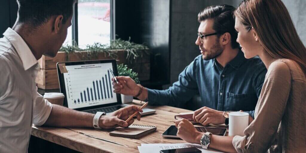 Business team reviewing financial charts on laptop screen with notebooks and coffee cups on wooden table.