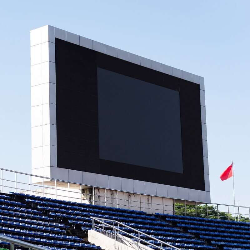 Large black digital display screen above blue stadium seating with flag pole and clear blue sky background.