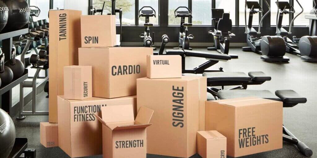 Cardboard boxes with fitness labels like "CARDIO" and "STRENGTH" stacked in gym with exercise equipment.