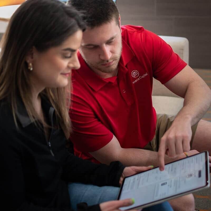 Man in red Geneva Capital polo shirt and woman reviewing documents on tablet together on couch.