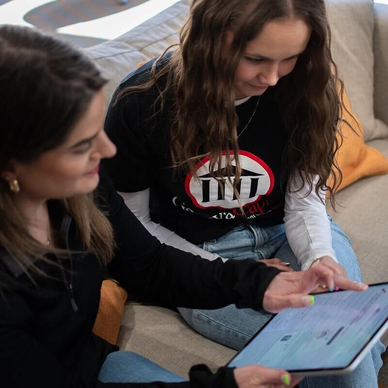 Two women sitting on couch looking at tablet together, one wearing Geneva Capital branded black shirt.