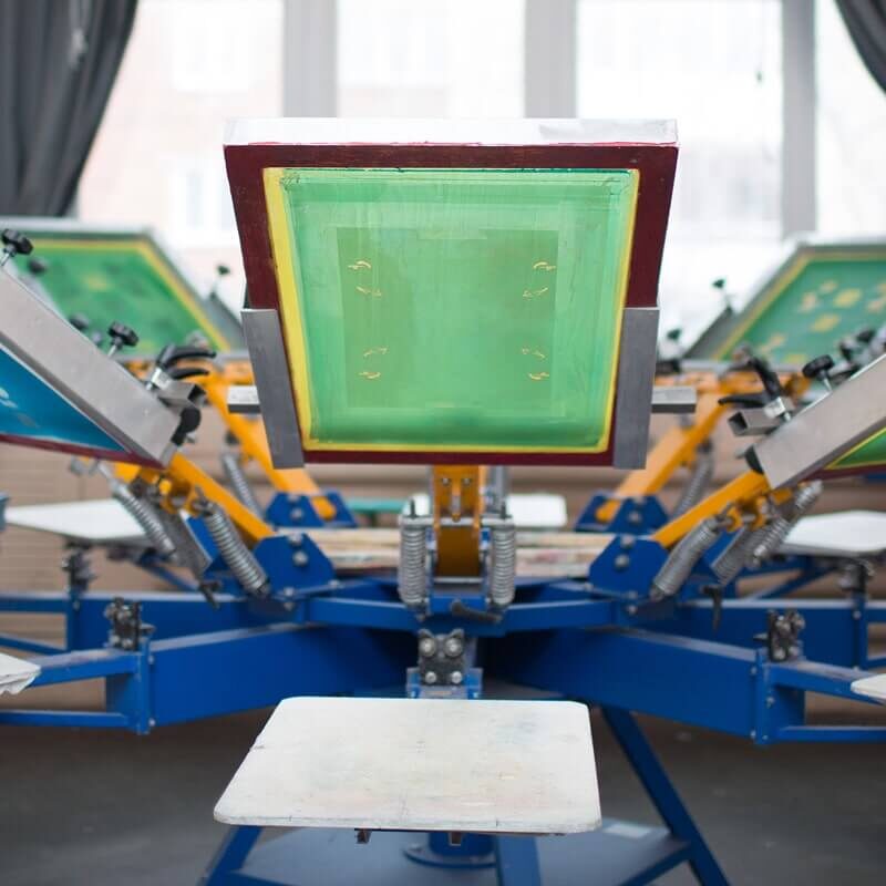 Industrial screen printing machine with blue frame, yellow arms, and green printing screens in manufacturing facility.