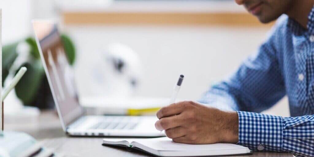 Hand resting on open notebook next to laptop computer on white desk for tax preparation work.