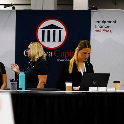 Two blonde women in business attire working at Geneva Capital booth with blue banner and laptops on white table.