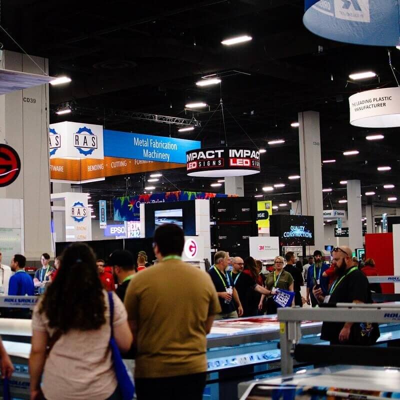 Crowded trade show hall with overhead banners for metal fabrication machinery, Impact Signs, and various industrial equipment vendors with attendees walking throughout.