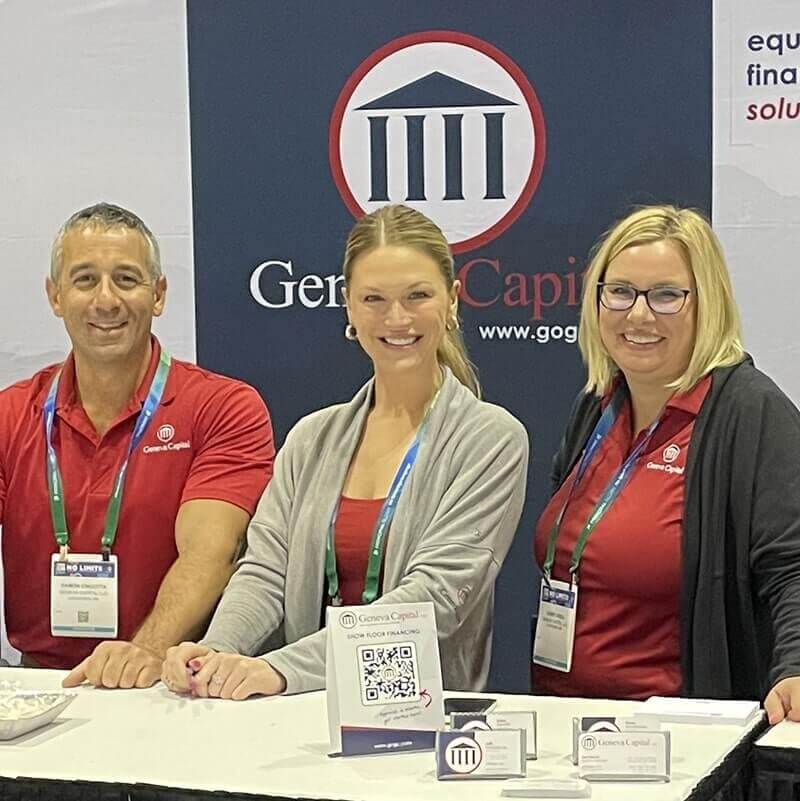 Three people posing at Geneva Capital booth - man in red polo, woman in gray cardigan, and woman in black jacket with red shirt, all wearing conference badges.